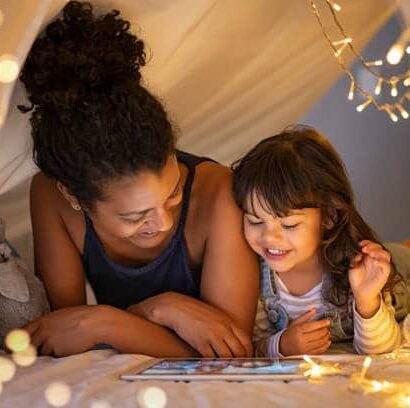 African mother and cute smiling girl using digital tablet while lying in illuminted tent in kid bedroom. Cheerful ethnic woman and lovely daughter on video call under a cozy hut. Lovely little girl with mom watching cartoon on digital tablet in bedroom.