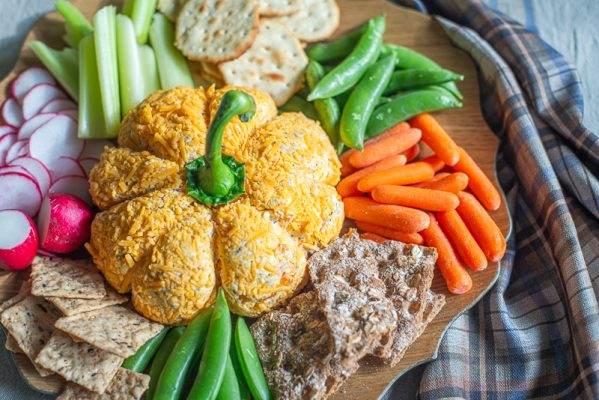 A pumpkin-shaped cheese ball surrounded by an assortment of crackers and fresh vegetables, including radishes, celery, sugar snap peas, and baby carrots, on a wooden board. The cheese ball is decorated with a green bell pepper stem, and a plaid cloth adds a cozy, autumnal touch to the presentation.