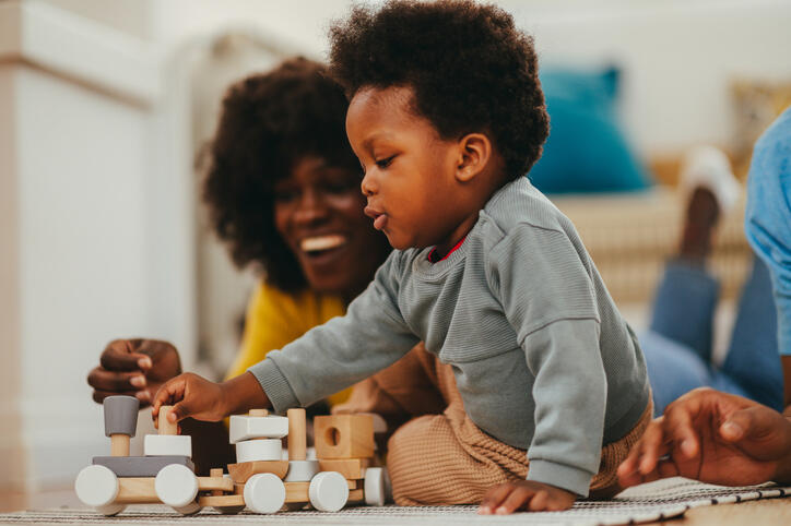 A cute little boy is playing and having fun with an educational Montessori toy while sitting on the floor at home. The parents are lying on the floor and smiling at their smart son.