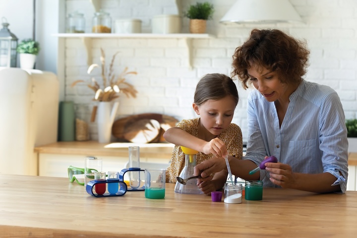 Concentrated small kid girl playing with young mom, involved in doing chemistry experiments together in modern kitchen. Happy mother educating little child daughter at home, science experiment