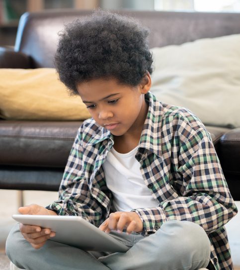 Serious elementary schoolboy of African ethnicity sitting by black leather couch on the floor in living-room and watching online educational video
