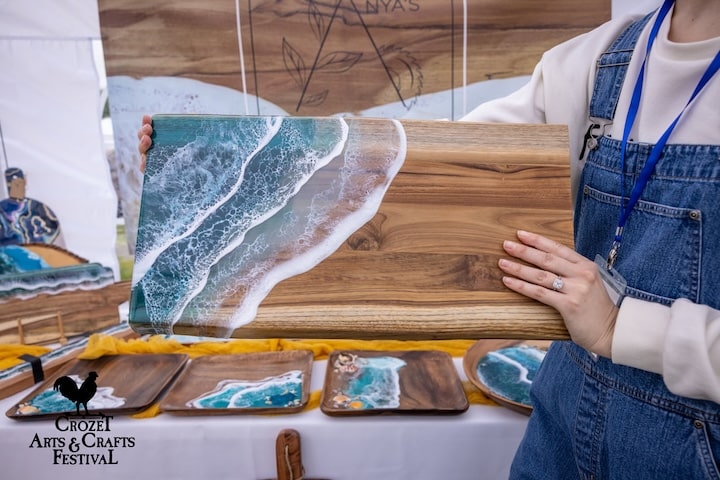 Artist in apron holding up wooden cutting board with ocean seafoam design on corner